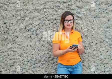 Portrait d'une femme millénaire, jeune caucasien portant des lunettes, un polo orange et un Jean appuyé contre un mur de calcaire tout en utilisant un téléphone Banque D'Images