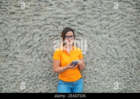 Jeune femme portant des lunettes, un polo jaune et un Jean bleu, se penchant contre un mur en pierre grise tout en souriant et regardant l'appareil photo Banque D'Images