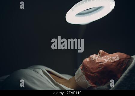 Alginate masque processus d'application. Une femelle mains d'une spatule de maintien d'esthéticienne. Femme couché sur un canapé dans le bureau du cosmetologiste. Soins du visage au salon de beauté. Banque D'Images