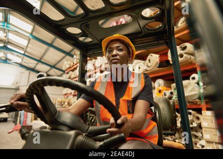 Jeune femme africaine opérateur de chariot élévateur roulant autour d'un entrepôt Banque D'Images