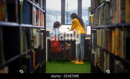 Bibliothèque de l'université : le garçon utilise un ordinateur personnel à son bureau, parle avec la femme de classe qui l'explique, l'aide avec l'attribution de classe. Étudiants ciblés Banque D'Images