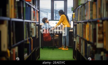 Bibliothèque de l'université : le garçon utilise un ordinateur personnel à son bureau, parle avec la femme de classe qui l'explique, l'aide avec l'attribution de classe. Étudiants ciblés Banque D'Images