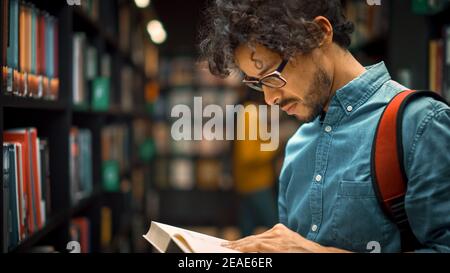 Bibliothèque de l'université : un garçon hispanique de talent portant des lunettes debout à côté de la bibliothèque lit le livre pour son affectation de classe et les préparatifs d'examen. Côté Banque D'Images