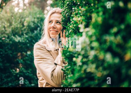 Portrait d'une femme blonde souriante dans un parc forestor vert en été portant un trench-coat beige tendance. Banque D'Images