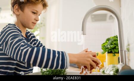 Dans la cuisine: Smart Little Boy préparation de la salade, lave Bell Pepper. Ses mains dans l'évier sous l'eau du robinet. Portrait de jeune garçon avec vue latérale Banque D'Images