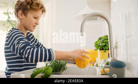 Dans la cuisine: Smart Little Boy préparation de la salade, lave Bell Pepper. Ses mains dans l'évier sous l'eau du robinet. Portrait de jeune garçon avec vue latérale Banque D'Images