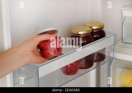 Jarres en verre avec confiture de framboise et de pomme sur l'étagère du réfrigérateur. Main femelle avec un pot de confiture maison rouge. Aliments végétariens sains fermentés. Banque D'Images