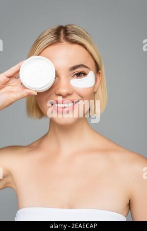 Femme blonde souriante dans le timbre oculaire tenant la crème cosmétique et isolé sur gris Banque D'Images