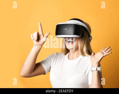 Une femme blonde heureuse en t-shirt blanc utilise des lunettes VR explore réalité augmentée en appuyant sur le bouton virtuel avec le doigt Banque D'Images