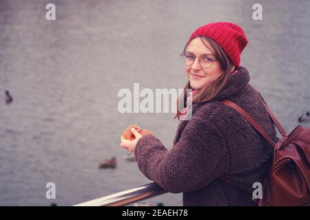 Une jeune femme en chapeau rouge nourrit les oiseaux sur le lac d'automne Banque D'Images