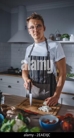 Photo d'une chemise blanche et d'un tablier de beau cuisinier tenant un couteau tranchant et posant sur un appareil photo dans une cuisine. Hipster Man en verres cuisine salade biologique Banque D'Images