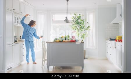 Jeune homme plein d'énergie et amusant avec des cheveux longs dansant dans la cuisine tout en portant un pyjama bleu. Cuisine moderne blanche et lumineuse avec vert sain Banque D'Images