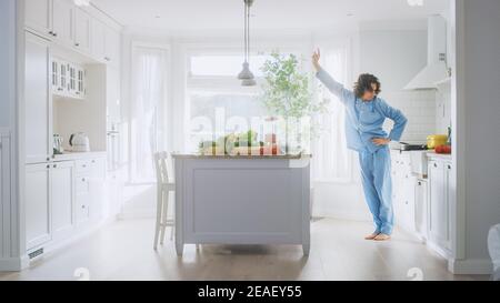 Fou drôle jeune homme avec long cheveux dansant dans la cuisine tout en portant Blue pyjama. Cuisine moderne blanche et lumineuse avec des aliments verts sains sur une Banque D'Images