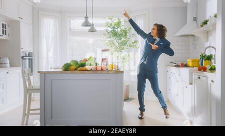 Jeune homme plein d'énergie et amusant avec des cheveux longs dansant dans la cuisine tout en portant un costume bleu. Cuisine moderne blanche et lumineuse avec vert sain Banque D'Images