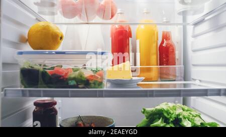 Point de vue POV prise de vue du réfrigérateur de cuisine. Réfrigérateur éclairé et plein de bons aliments : tomates cerises, fromage, jus de fruits frais et chou, salade faite Banque D'Images