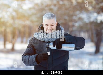 Joyeux homme âgé dans une veste chaude et un foulard versant le thé de thermos ballon dans un parc d'hiver enneigé. Activités saisonnières Banque D'Images