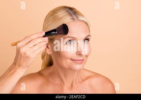 Portrait en gros plan d'une charmante femme d'âge moyen appliquant de la poudre naturelle tonalité de correction isolée sur fond beige pastel Banque D'Images