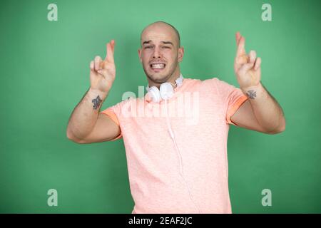 Jeune homme chauve utilisant un casque sur fond vert gesturant le doigt traversé souriant avec espoir et aspect Banque D'Images