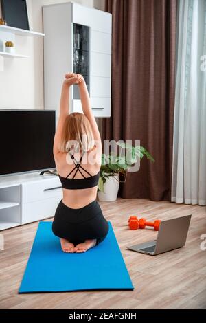 Femme sportive faisant du yoga de forme physique à la maison via un ordinateur portable par appel vidéo en ligne à distance. Jeune femme perdant du poids par l'entraînement en ligne à distance de gym. Vue arrière Banque D'Images