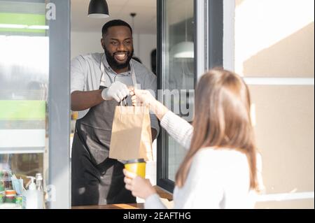 Un serveur afro-américain amical et souriant sert une cliente féminine, un gérant de café multiracial tient une commande à emporter à emporter Banque D'Images