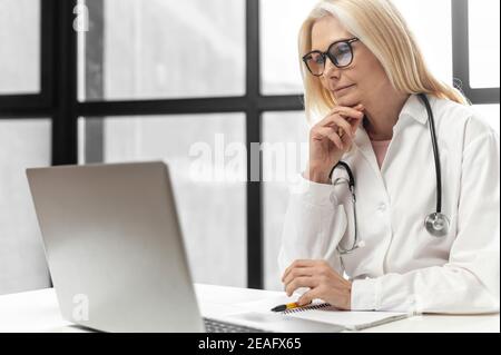 Une femme mature blonde médecin portant des lunettes, un manteau médical et un stéthoscope regardant un séminaire en ligne, assis à son bureau avec l'ordinateur portable Banque D'Images