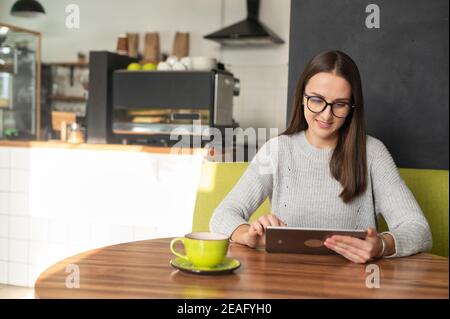 Femme souriante portant des lunettes élégantes à l'aide d'une tablette numérique pour faire défiler l'onglet des actualités, répondre à des e-mails, surfer sur le Web assis à table avec une tasse Banque D'Images