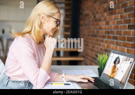 Femme d'affaires blonde mûre intelligente dans des lunettes, assise à la maison, étudiant, prenant des cours virtuels, ayant une réunion en ligne sur un ordinateur portable Banque D'Images