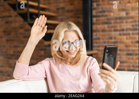 Femme mûre souriante dame blonde parlant en ligne sur le téléphone mobile, participant à une conférence, assis sur le confortable canapé vidéoconférence avec le Banque D'Images