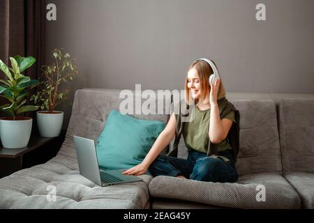 Une jeune femme heureuse est assise sur un canapé avec un ordinateur portable dans un casque. Travail à distance, étude en ligne ou achats en ligne. Reposez-vous à l'intérieur de la salle de séjour Banque D'Images