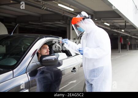 Site d'essai du drive-in COVID-19, personnel médical portant un EPI et un écran facial en train d'effectuer un essaiing nasal, jeune homme conducteur patient attendant assis dans un automate Banque D'Images