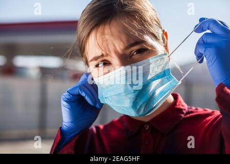 Une jeune femme inquiet a mis un masque de protection individuelle sur le visage/l'a éteint en public, portant des gants chirurgicaux de protection en latex bleu, regardant la caméra avec stress et un Banque D'Images