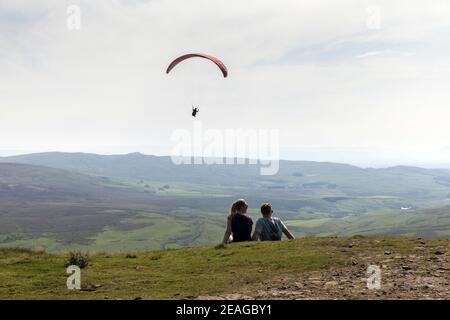 Un couple s'assoit au sommet de Pen-y-ghent dans le Yorkshire Dales en tant que parapente vole au-delà Banque D'Images