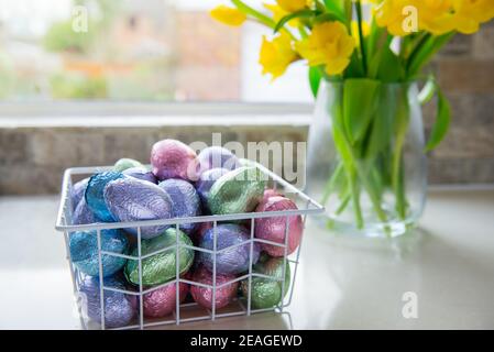 Panier en métal avec œufs de pâques au chocolat en feuilles de couleur et bouquet de tulipes jaunes et de jonquilles fleurissent sur une table de cuisine blanche près de la fenêtre. Festiv Banque D'Images