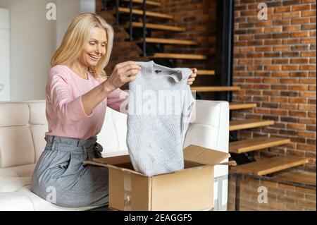 Une femme blonde élégante et satisfaite ouvre une boîte de carton, déballant un cadeau reçu, se sentant curieux de l'article commandé dans un magasin en ligne Banque D'Images