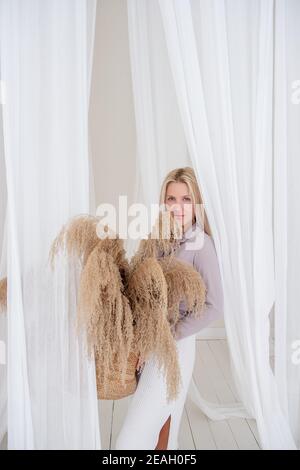 Jeune femme blonde dans une chemise lilas, robe blanche, tient dans ses mains panier de paille avec l'herbe de pampas parmi les aérées, développant des tissus de tulle. Minimali Banque D'Images