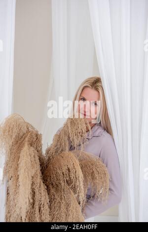 Jeune femme blonde dans une chemise lilas, robe blanche, tient dans ses mains panier de paille avec l'herbe de pampas parmi les aérées, développant des tissus de tulle. Minimali Banque D'Images