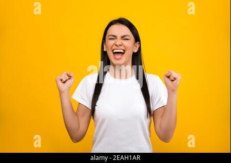 Heureuse joyeuse fille caucasienne portant un t-shirt blanc de base, se réjouissant dans le succès ou la victoire, debout sur fond bleu isolé Banque D'Images