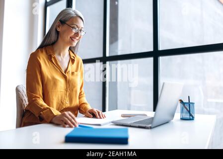 Une femme d'affaires asiatique mûre satisfaite, vêtue de lunettes élégantes et décontractées, utilise un ordinateur portable pour le travail à distance, l'apprentissage à distance, l'enseignement en ligne Banque D'Images