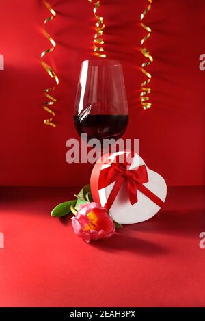 Verre de vin, boîte cadeau en forme de coeur, tulipe fraîche et rubans dorés sur fond rouge. Amour célébration romantique de la Saint-Valentin, le 14 février Banque D'Images