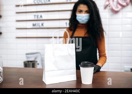 Café à emporter. Du café ou du thé dans un verre jetable en carton et un sac en papier avec dessert sont sur le comptoir du bar, à l'arrière-plan se trouve le barista ou la serveuse défoqué dans un masque médical Banque D'Images