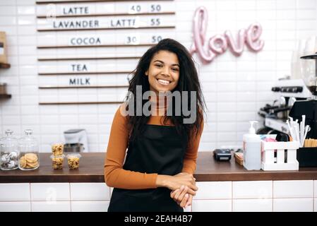 Concept de propriétaire d'entreprise de café. Jeune barista afro-américain ou serveuse en tablier noir souriant à l'appareil photo près du comptoir du café. Banque D'Images