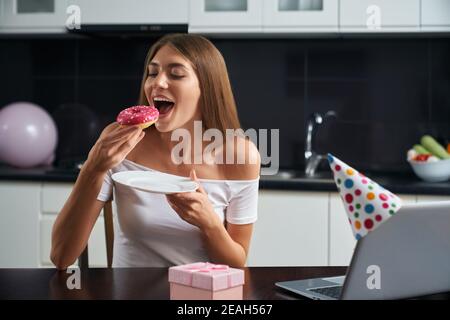 Une jeune femme satisfaite mangeant un délicieux donut coloré tout en célébrant son anniversaire par appel vidéo sur ordinateur portable. Concept d'isolement et tec moderne Banque D'Images