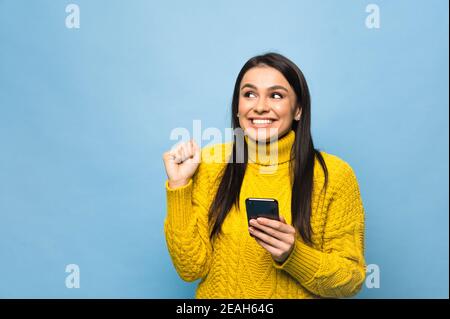 Portrait d'une jeune femme du caucase heureuse, tient le smartphone à portée de main, se réjouit d'une bonne nouvelle ou a reçu un message inattendu, se tient sur un fond bleu isolé Banque D'Images