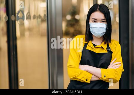 Une jeune femme asiatique serveuse ou propriétaire d'un restaurant, d'un café ou d'un magasin dans un masque protecteur et un tablier se tient à l'entrée. Petite entreprise dans le contexte de la pandémie du coronavirus Banque D'Images