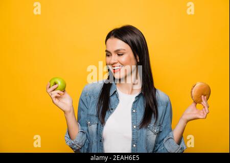 Restauration rapide ou alimentation saine. Caucasien belle fille brune tient une pomme dans une main et dans l'autre hamburger, faisant un choix, debout sur un fond isolé orange Banque D'Images