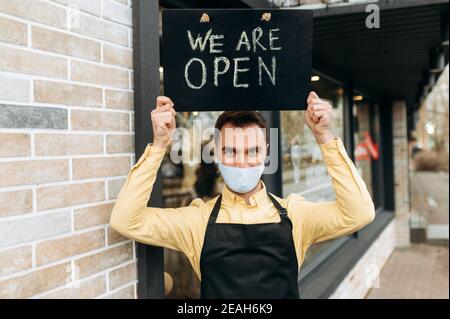 Bienvenue, nous sommes ouverts. Le serveur mâle caucasien se tient à l'extérieur près d'un restaurant ou d'un café portant un masque médical de protection et un tablier noir et tient le panneau OUVERT au-dessus de la tête. Prendre en charge le concept de petite entreprise Banque D'Images