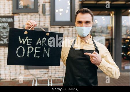 Bienvenue, nous sommes ouverts. Le serveur mâle caucasien se tient à l'extérieur près d'un restaurant ou d'un café portant un masque médical de protection et un tablier noir et tient le panneau OUVERT. Prendre en charge le concept de petite entreprise Banque D'Images