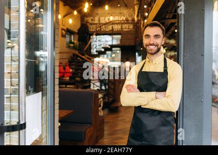 Portrait d'un serveur, barista ou propriétaire d'un restaurant, d'un café ou d'un bar à l'entrée portant un masque de protection et un tablier de travail noir avec bras croisés et Banque D'Images