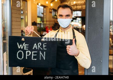 Nous sommes OUVERTS. Portrait d'un serveur ou barista caucasien satisfait debout à l'entrée du restaurant ou du café, portant un masque médical sur son visage et son tablier noir. Employé montrant le panneau OUVERT dans son Banque D'Images