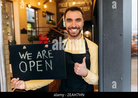 Bienvenue, nous sommes ouverts. Portrait du serveur, du barista ou du propriétaire caucasien, joyeux et masculin, debout à la porte d'un restaurant ou d'un café, tient un panneau OUVERT, et un sourire amical. Prendre en charge le concept de petite entreprise Banque D'Images